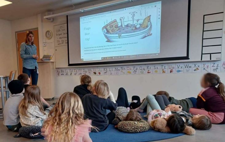 En grupp barn sitter på golvet och lyssnar på en kvinna som talar och visar en bild på en stor skärm.