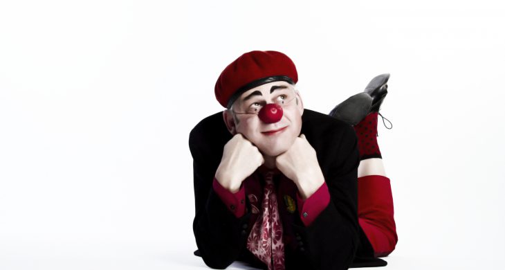 En clown som ligger på mage med benen upp och hakan mot händerna. Han har på sig en rund clownnäsa.