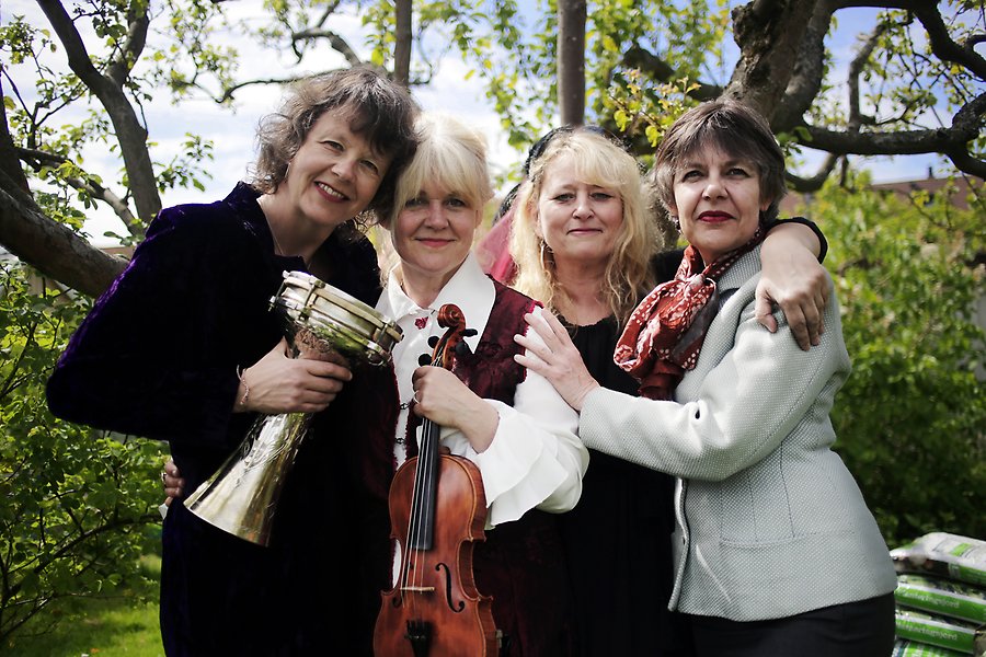 Fyra kvinnor står tätt tillsammans och ler in i kameran. En håller en bongoliknande trumma och en annan en fiol. I bakgrunden syns en trädgård med träd.