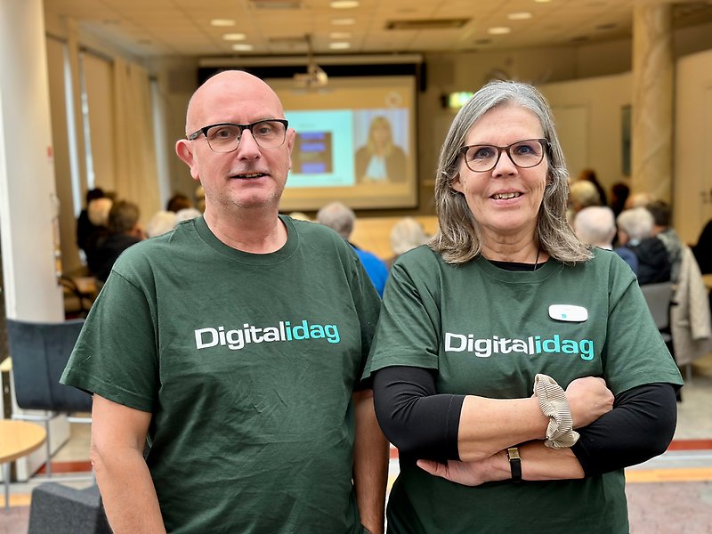 Två glada personer med mörkgröna t-shirts där det står Digital idag på. I bakgrunden syns en webbföreläsning med flera personer i publiken.