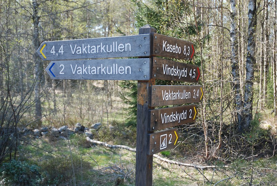 Foto på en stolpe med skyltar som hänvisar till olika vandringsleder. Skyltarna visar även ledernas längd och är placerad i skogsmiljö.