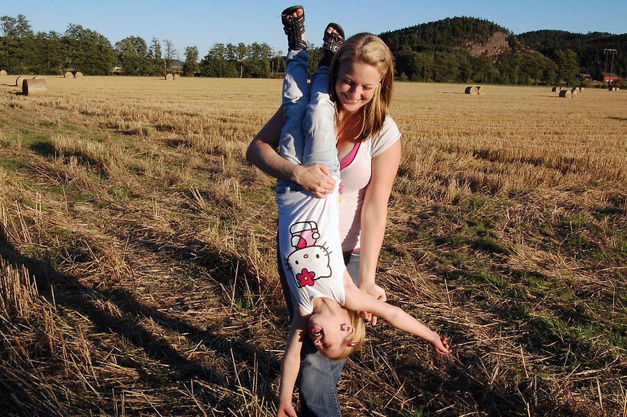 Foto på en kvinna som håller ett barn upp och ner medans båda två skrattar. I bakgrunden syns en gul åker och ett berg.