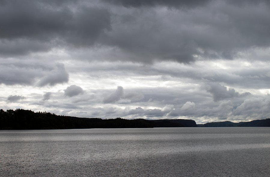 Foto på en sjö. Mörka moln hänger över sjön med krusningar på ytan. Vid horisonten syns skog och berg..