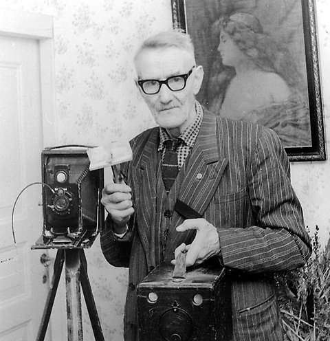 Svartvitt äldre foto på en äldre man i randig kavaj, slips och glasögon. Bredvid honom står en äldre kamera på stativ. I handen håller han en låda. I bakgrunden syns en tavla och en dörr.