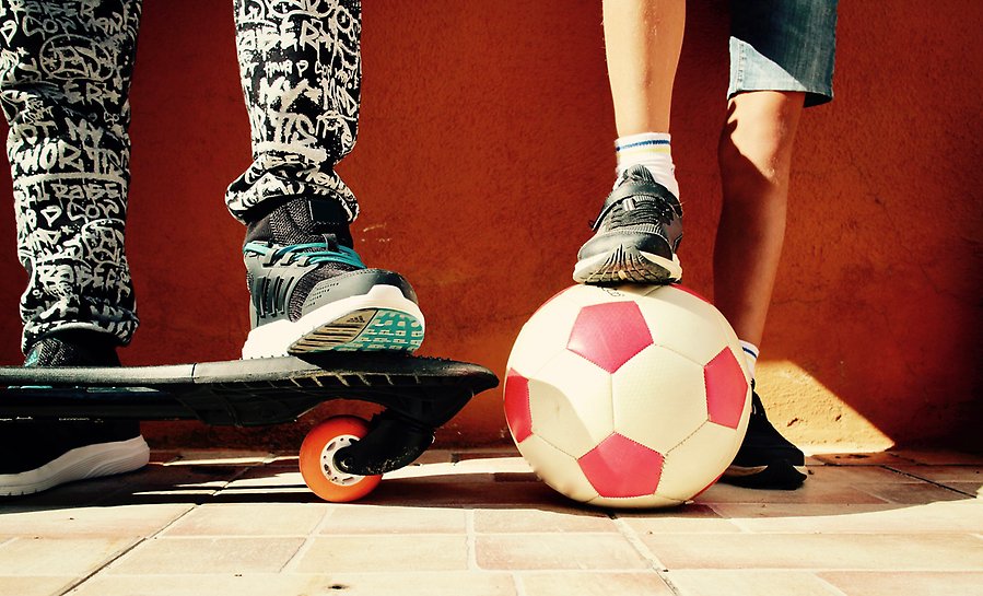 Närbild på två barns ben och fötter. De har träningsskor på sig, den ena har en fot på en skateboard och den andre har en fot på en fotboll.