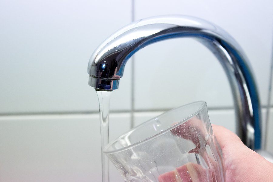 Närbild på en hand som håller upp ett vattenglas under en rinnande vattenkran. I bakgrunden syns vitt kakel.