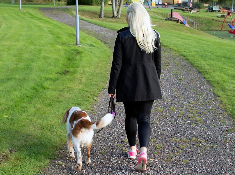 Foto på en kvinna med blont hår och svarta kläder som går på en grusväg i en park med en vit-röd hund i koppel bredvid sig.