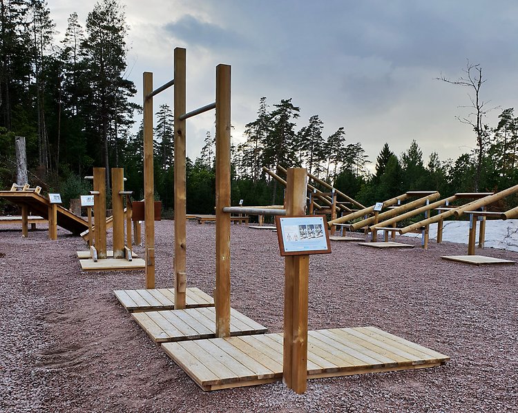 Foto på ett utegym med olika stationer gjorda av trä som står på en grusplan. I bakgrunden syns skog och en molnig himmel. 