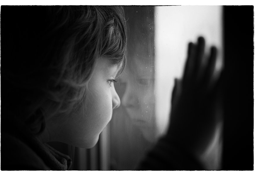 Svartvit bild där ett litet barn står nära ett fönster, håller en hand mot det och tittar ut.