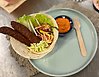 En vegetarisk maträtt med grönsaker och två avlånga biffar i en tortilla-wrap. Bredvid på tallriken ligger en trägaffel och en liten skål med orange ajvar-gurt.