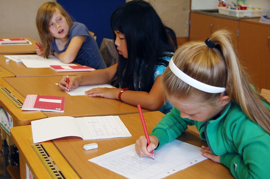 Bild på tre barn som sitter vid skolbänkar och skriver.