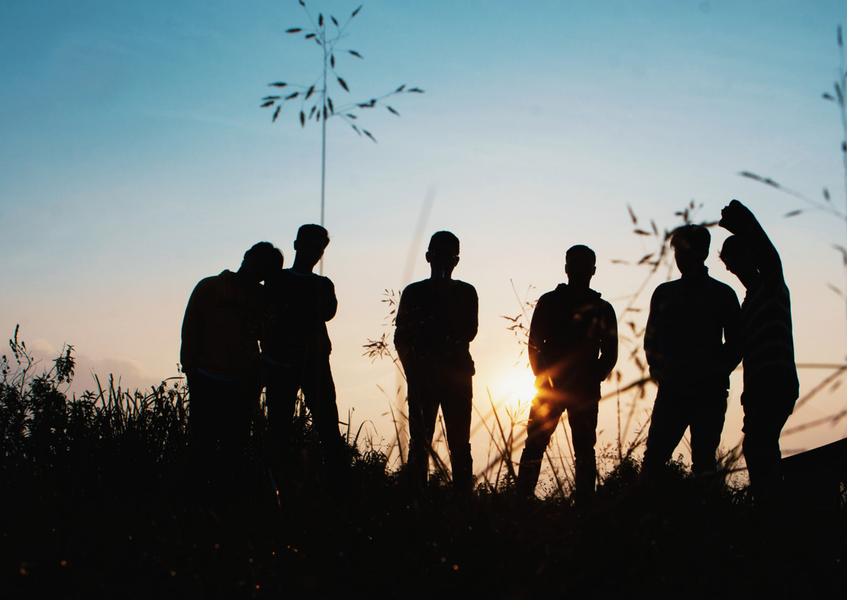 I silhuett mot en lågt stående sol syns sex stycken ungdomar som står i högt gräs.