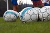 Foto på flera fotbollar som ligger på grönt gräs. I bakgrunden syns ben på spelare.