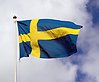 Foto på en vajande svensk flagga på en flaggstång. Flaggan har ett gult kors på en mörkblå botten. Moln syns på den blå himlen.