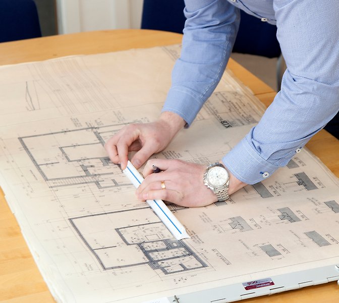 Foto på en en persons armar som lutar sig över ett skrivbord och mäter med linjal på ett papper.