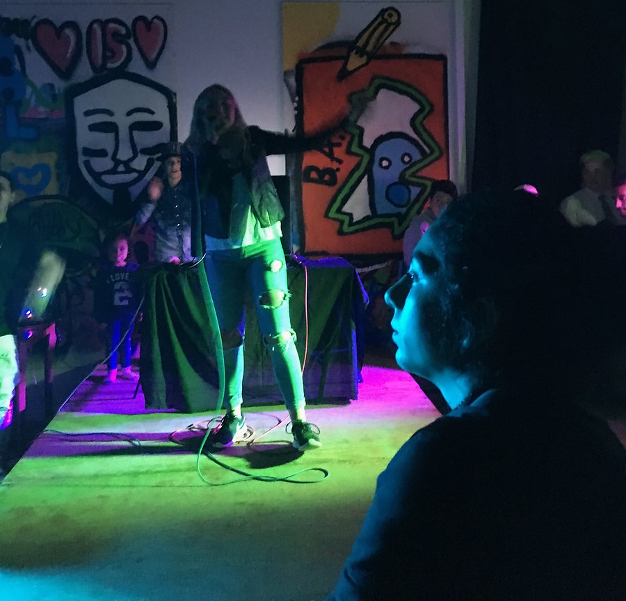 En yngre tjej står på en scen och sjunger. Ljuset är grönt, blått och lila. Bakom scenen syns en grafittiväg. I förgrunden syns profilen av en tjej i publiken.