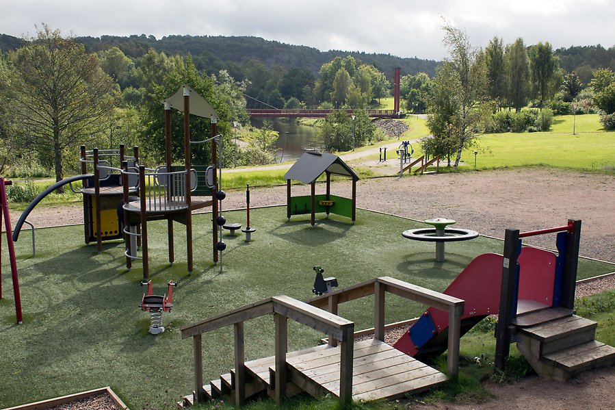 Bild över en park med stora grönytor och vatten, och en lekplats i förgrunden.