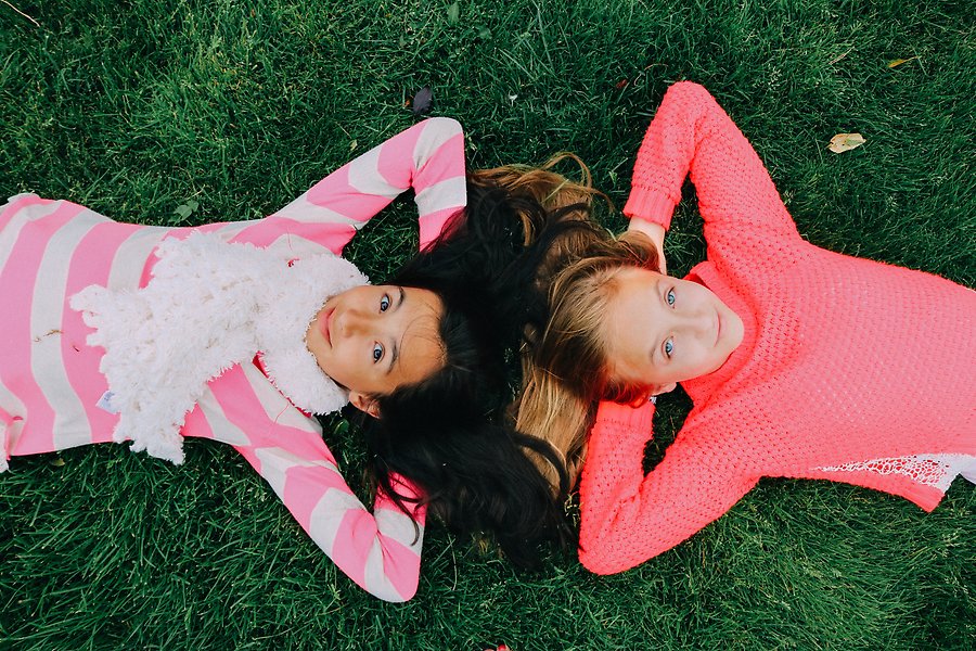 Foto på två flickor som ligger på rygg i gräset, huvud mot huvud. Färgglada tröjor.