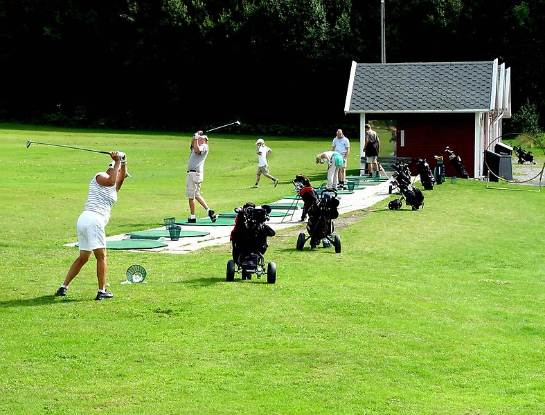 Foto över en drivingrange i sommartid där flera personer svingar golfklubbor.