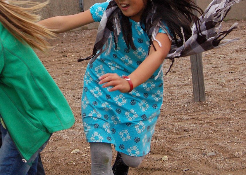 Foto på två flickor som springer på sand på en lekplats. Den ena flickan har mörkblont hår och grön tröja, den andra flickan har mörkbrunt hår och blå klänning.