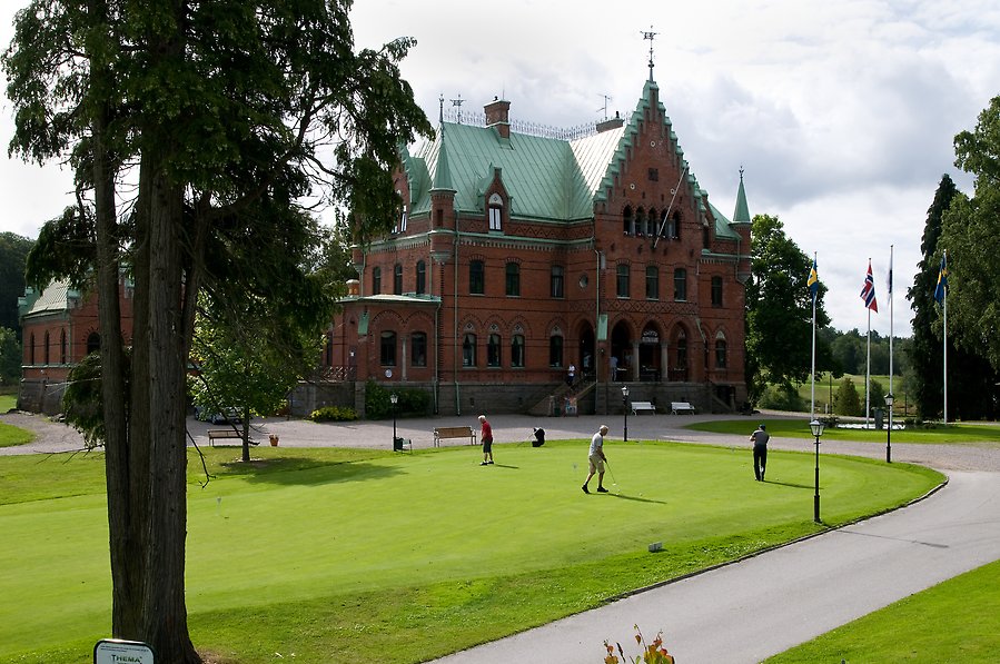 Foto på ett slott i tegel med tinnar och torn, tak av grönärgad koppar. I förgrunden syns träd och en golfbana där några personer spelar golf.