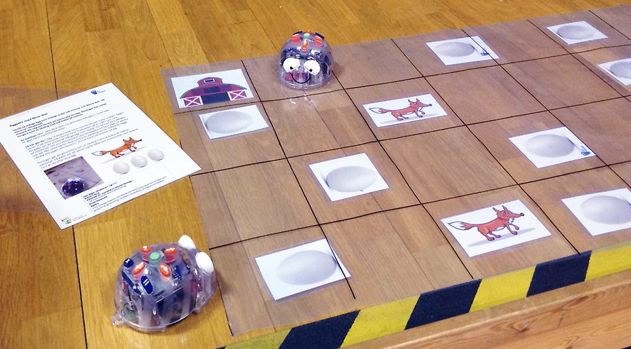 På ett bord ligger ett genomskinligt ark indelat i rutor. I vissa av rutorna ligger kort med olika figurer på. På bordet stör också två små robotar i plast som är blå till färgen.