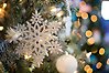 I en julgran hänger en vit snöflinga. Längre bort i bilden syns en vit julgranskula samt ljus.