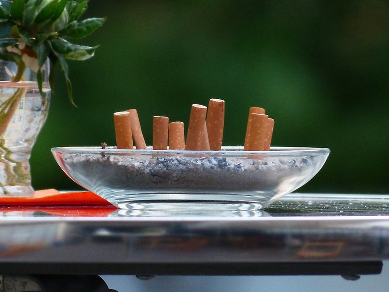 Foto på ett askfat med många fimpad cigaretter nerstuckna rakt upp och ned. Faten står på ett bord utomhus med en vas med blommor i bredvid.