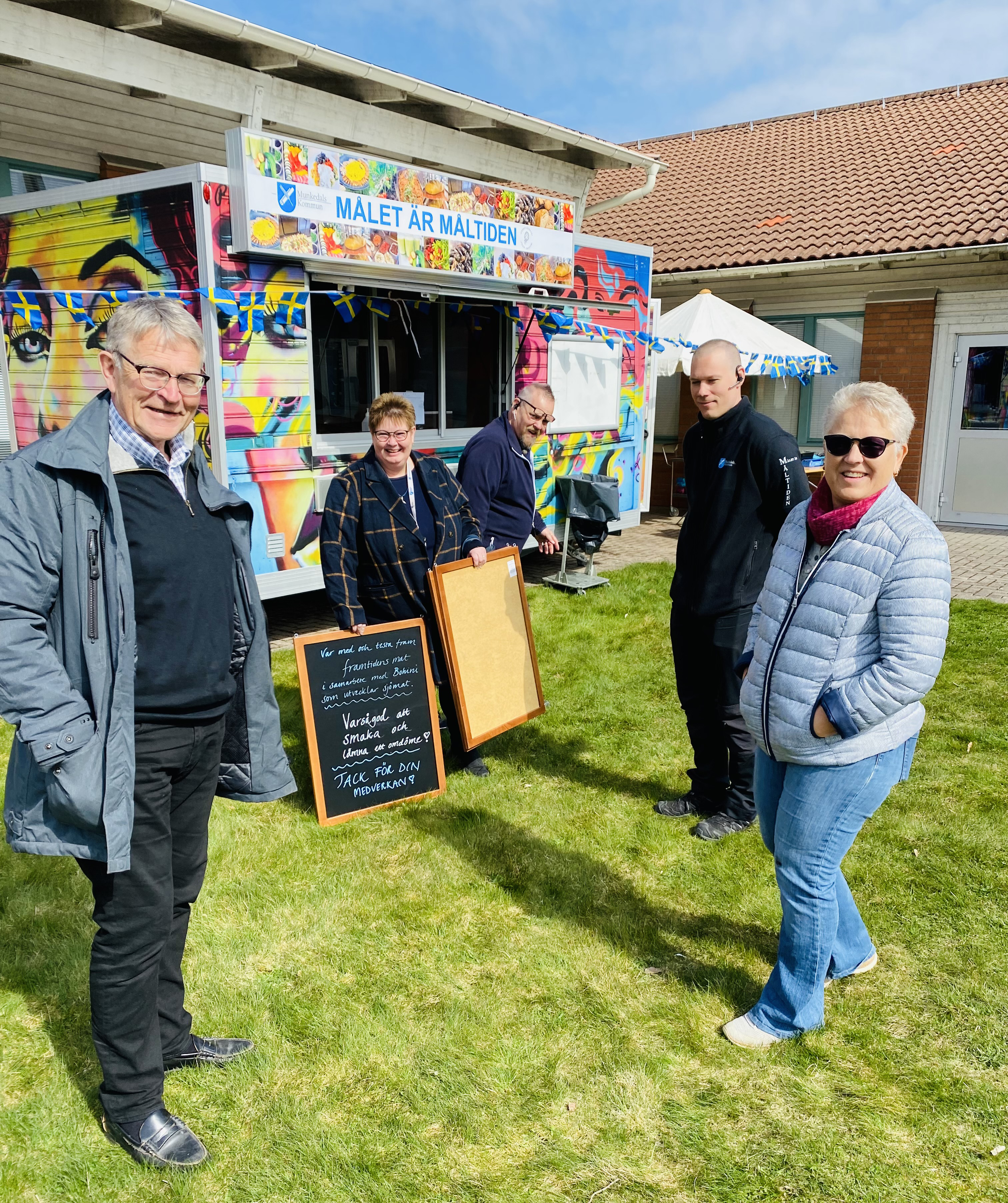 I strålande solsken står fem personer framför foodtrucken. Två av dem håller i handskrivna griffeltavelskyltar som de är på väg att sätta upp. Foodtrucken är målad i glada färger. På foodtrucken står det Målet är måltiden tillsammans med Munkedals kommuns logga och loggan för Foodfluencers. 