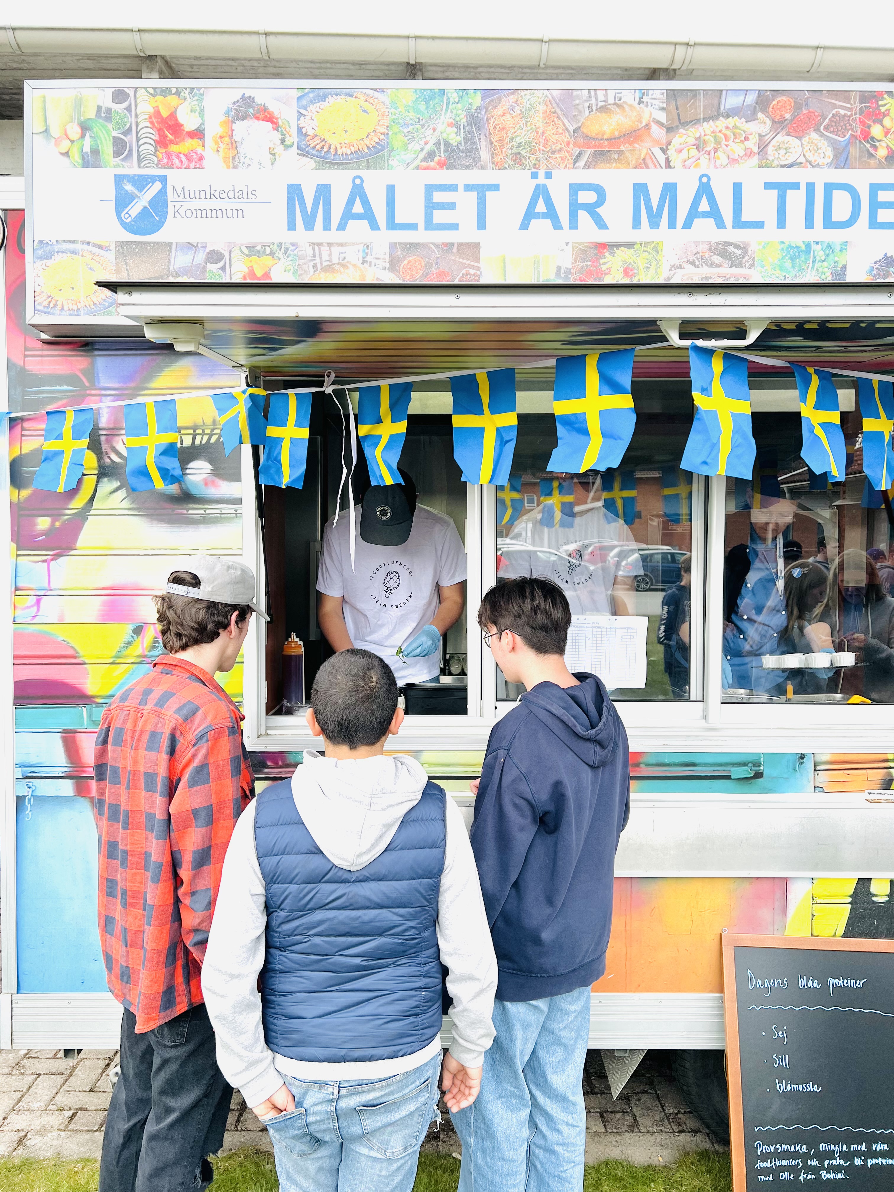 Tre ungdomar står framför foodtrucken och håller på att bli serverade. Foodtrucken är målad i glada färger. På foodtrucken står det Målet är måltiden tillsammans med Munkedals kommuns logga.