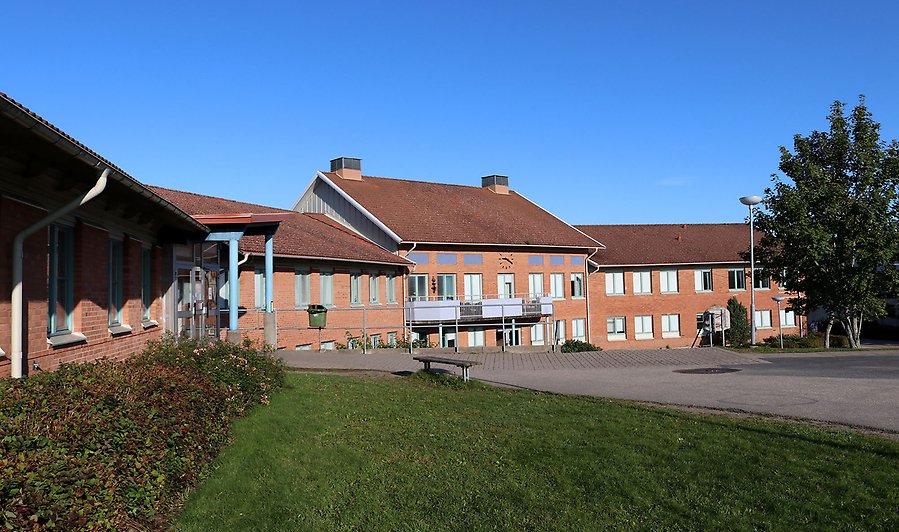 Stor tegelbyggnad en solig dag. Runt om gräs och träd. 