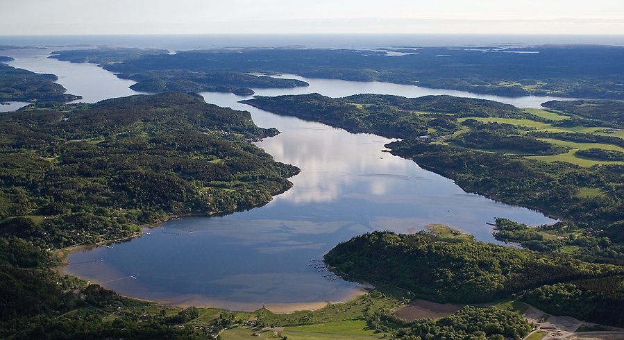 Flygfoto från inlandet ut mot kusten. i förgrunden syns en vik. Två fjordar möter sedan varandra innan de tillsammans mynnar ut i havet. På land ses skogs- och jordbrukslandskap.