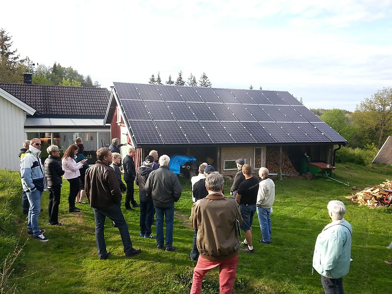 En samling av personer står på en gräsmatta utomhus vid en  röd byggnad med svart tak med solceller på taket.