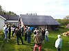 En samling av personer står på en gräsmatta utomhus vid en  röd byggnad med svart tak med solceller på taket.