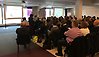 En samling på 70 personer sitter i stolar och lyssnar på en person som berättar om säkerhetsskydd vid en duk där en presentation projiceras.