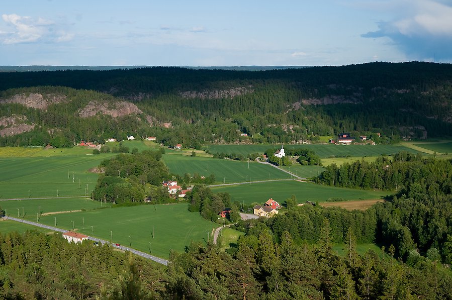 Vy över en grön jordbruksbygd i en dal. Mitt i bilden ligger en kyrka. Här och var syns små gårdar.