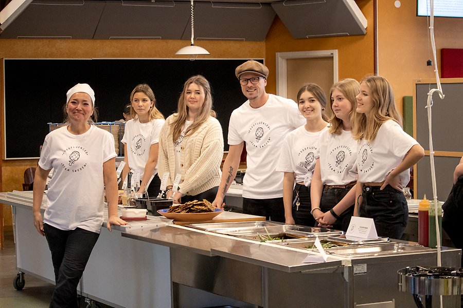 Två vuxna och fem ungdomar personer står vid en matservering iklädda vita t-shirts