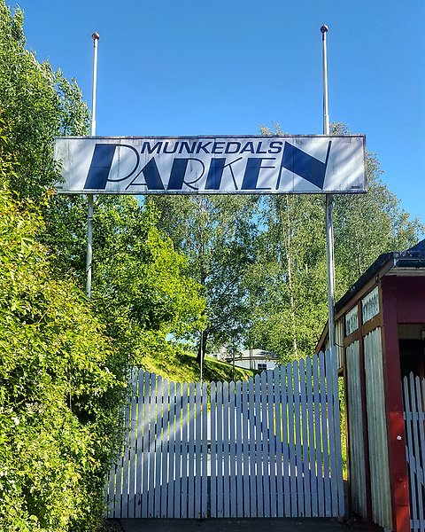 Ingången till Munkedals folkpark. Entrén har vita grindar av trä och en vit skylt med blå text där det står: Munkedalsparken. Bilden är tagen på sommaren en solig dag och grönska omger entrén..