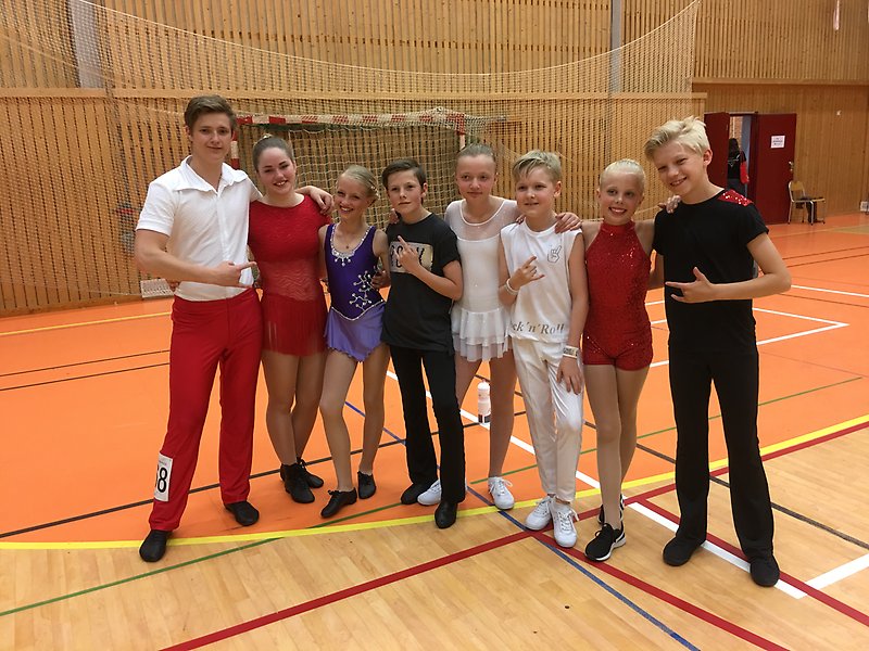 En grupp av barn och tonåringar står tillsammans på rad och ler in i kameran. De står inne i en gymnastiksal och har tävlingskläder för dans på sig.