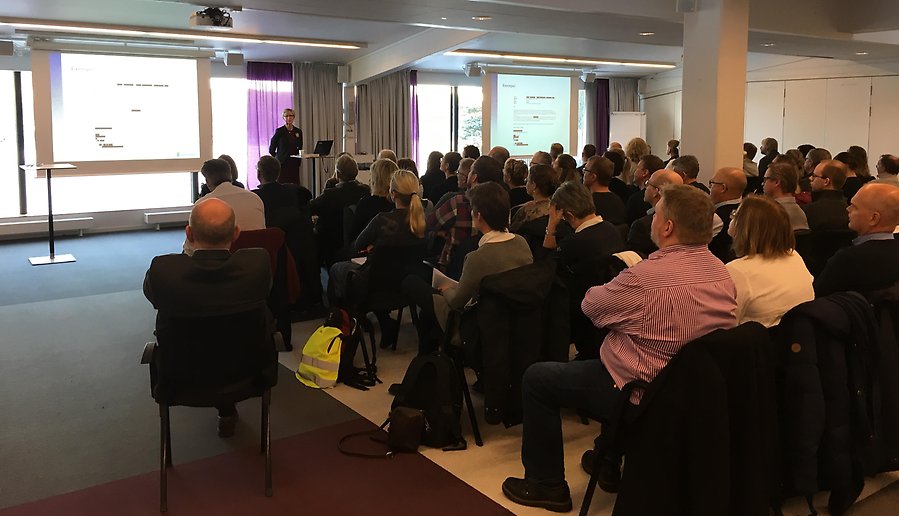 En samling på 70 personer sitter i stolar och lyssnar på en person som berättar om säkerhetsskydd vid en duk där en presentation projiceras.